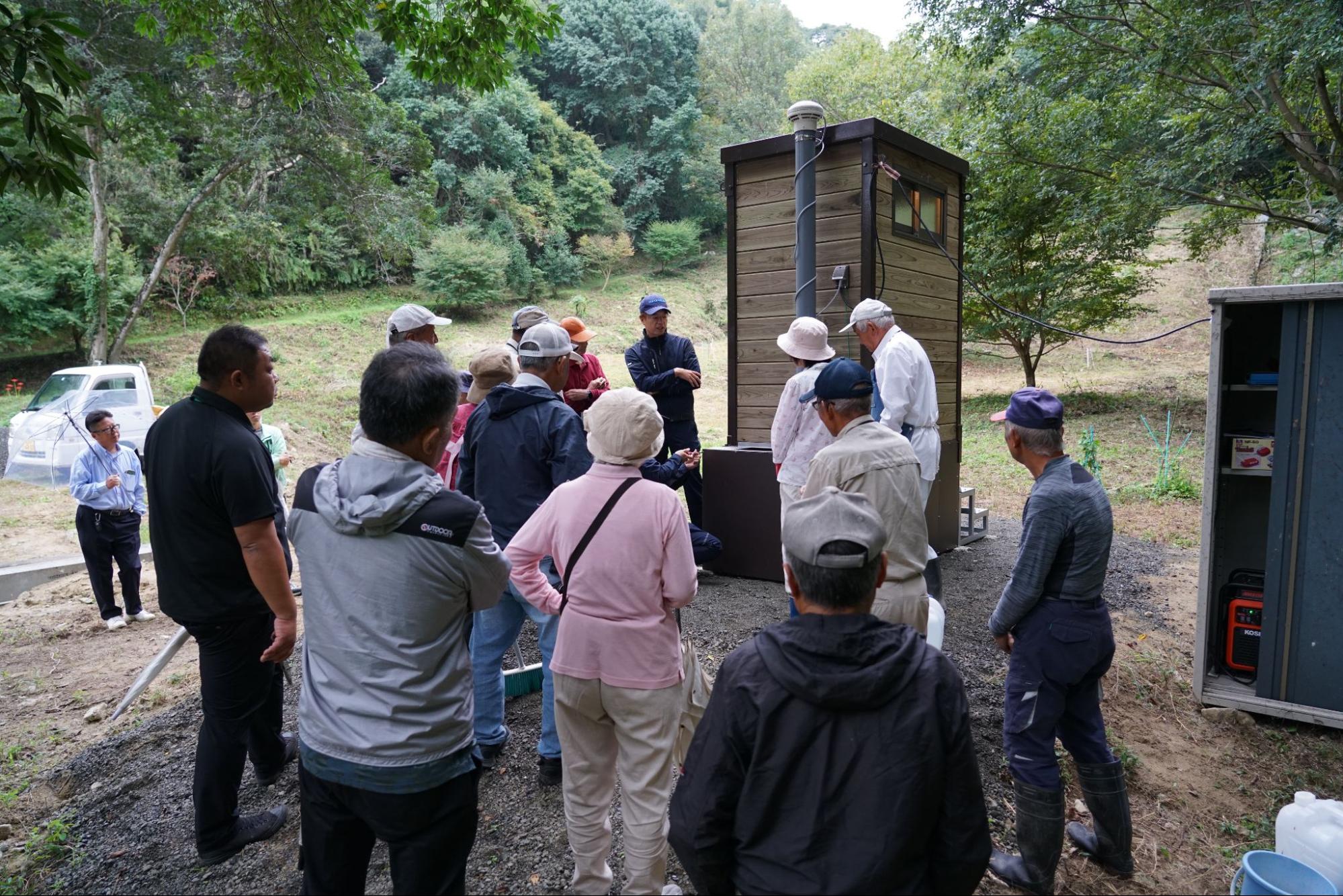 バイオトイレの完成披露会で説明を受ける地域住民と、周囲の設備を確認する様子