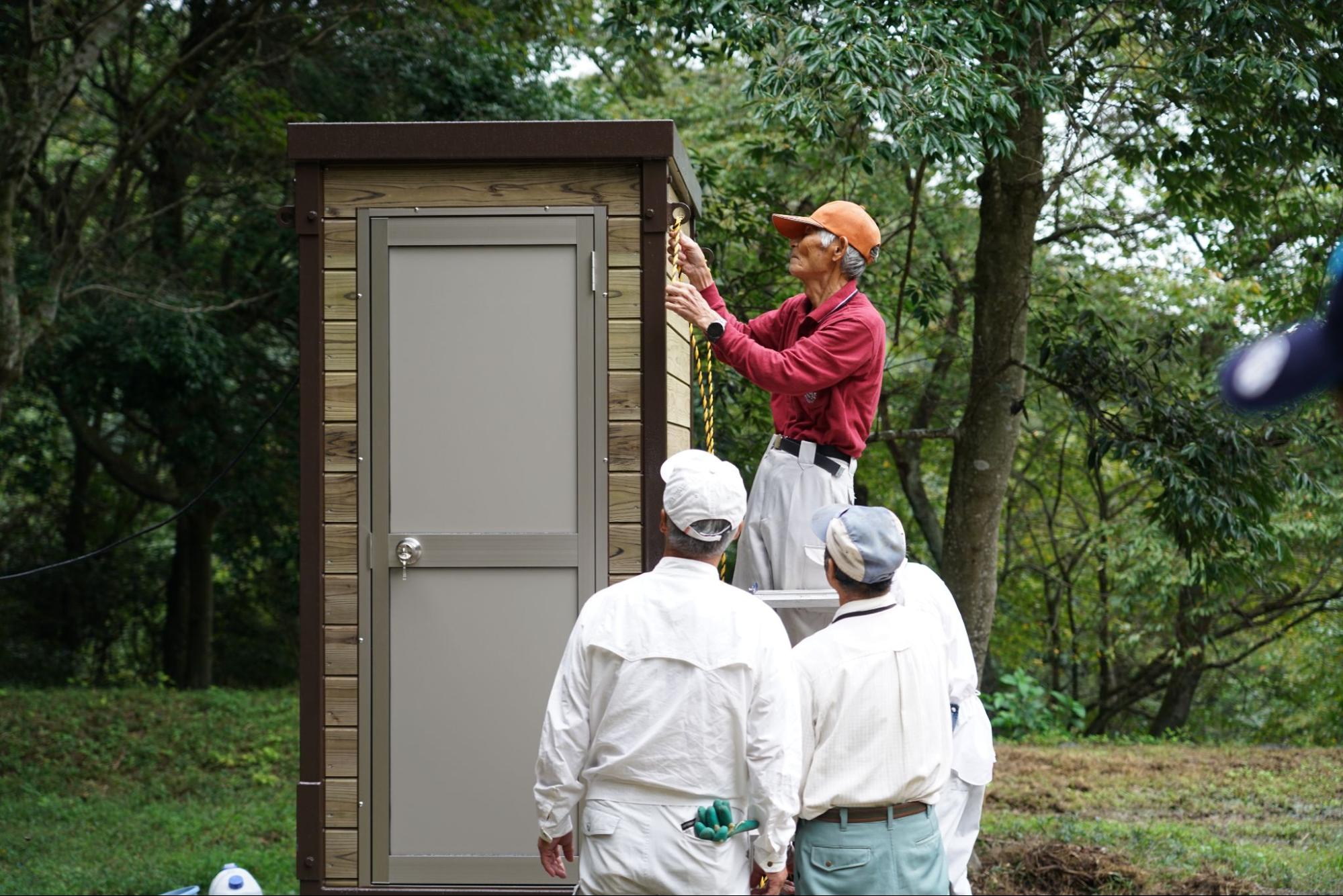 新設されたバイオトイレの外壁にロープで屋根補強を施す地域住民の男性と見守る作業